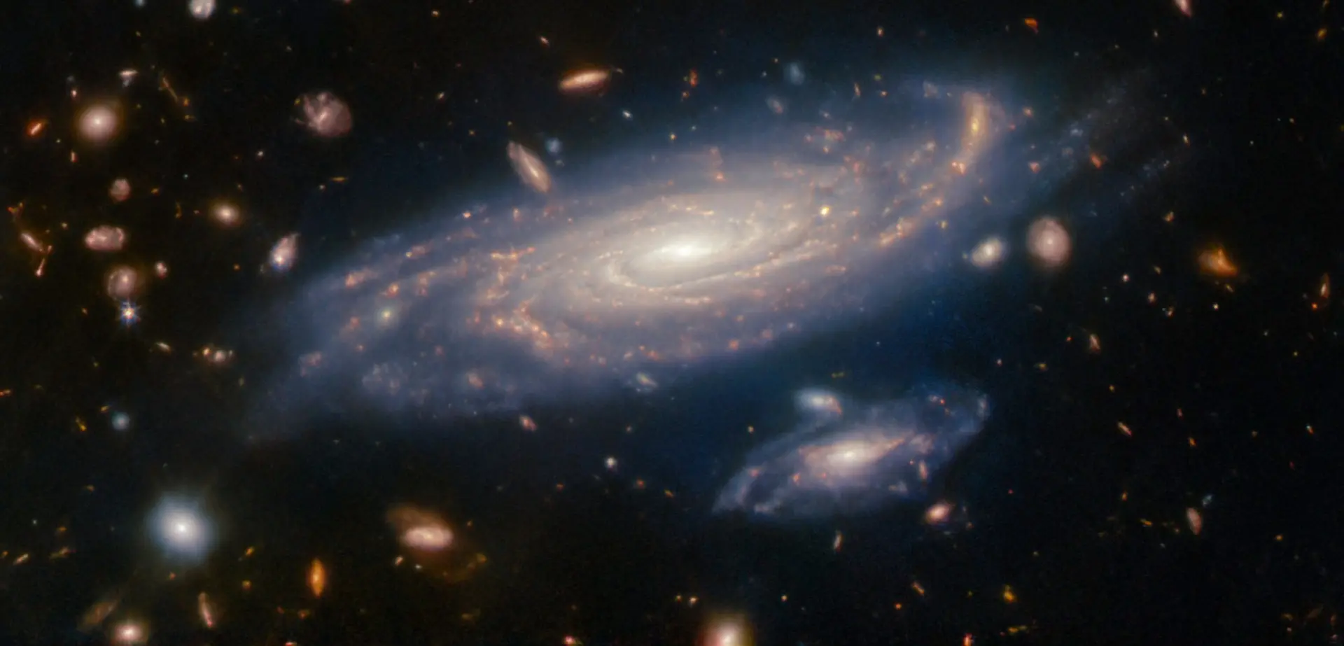 Foto de duas galáxias em close, mais esbranquiçadas, e milhares aos fundo, alaranjadas, obtidas pelo Telescópio Espacial James Webb
