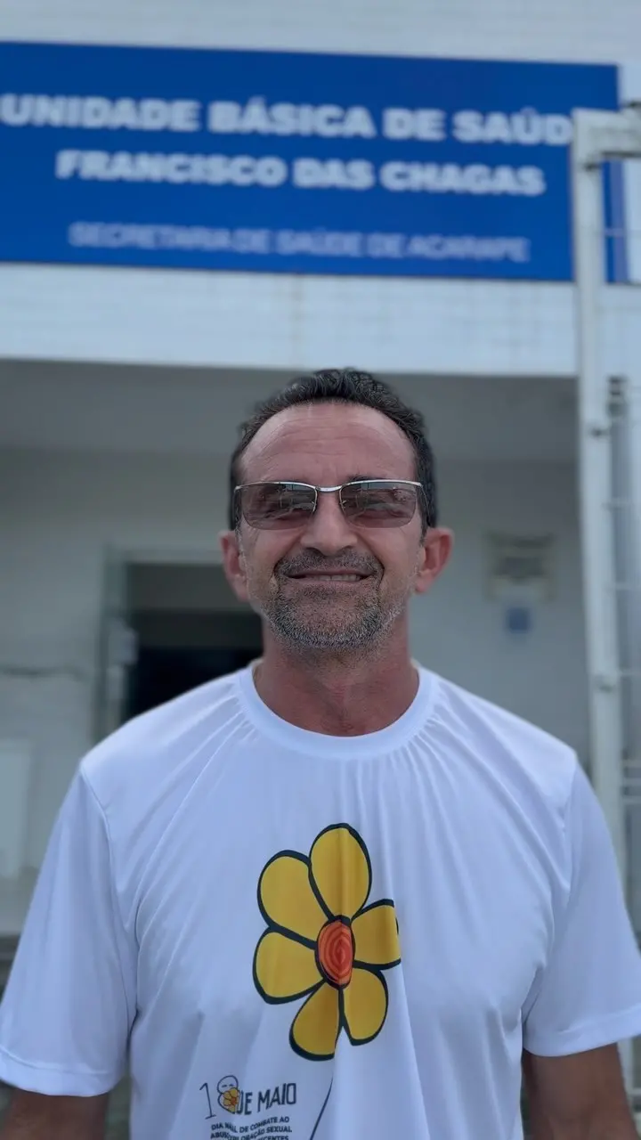 Homem sorridente com óculos escuros, usando camiseta branca com símbolo de uma flor amarela e um centro laranja, em frente a um hospital com banner azul ao fundo promovendo a saúde básica.