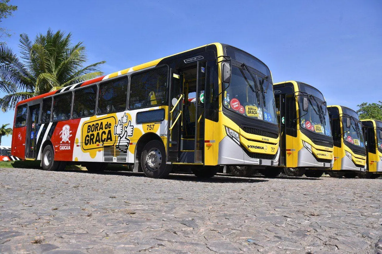 Imagem mostra quatro ônibus estacionados em pátio da Prefeitura de Caucaia. Os veículos têm as cores vermelha, amarela e branca, e mostra o letreiro Bora de Graça, nome de programa de passe-livre municipal.