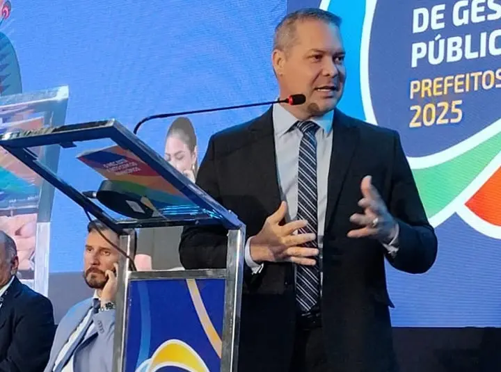 A imagem mostra Heitor Freire de terno escuro, camisa clara e gravata listrada falando ao microfone no palco do Seminário dos Prefeitos. Ele está em pé atrás de um púlpito transparente com detalhes em metal e uma placa colorida que diz: 