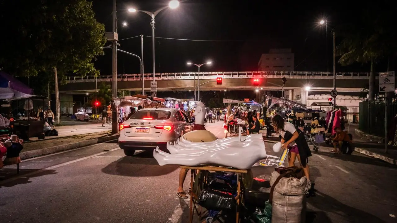Foto que contém um táxi trafegando em meio a barracas de roupas na avenida Alberto Nepomuceno durante a Feira da Madrugada