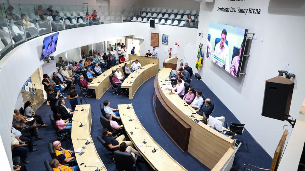 Imagem mostra o plenário da Câmara Municipal de Juazeiro do Norte durante uma sessão, com vereadores sentados em mesas em formato semicircular ao centro e público assistindo nas galerias laterais e superiores. Um grande telão exibe a imagem do prefeito Glêdson Bezerra falando ao microfone, durante visita à Casa. Na parede ao fundo está escrito “PLENÁRIO Presidenta Dra. Yanny Brena”. A sala é bem iluminada, com bandeiras ao fundo e estrutura moderna.