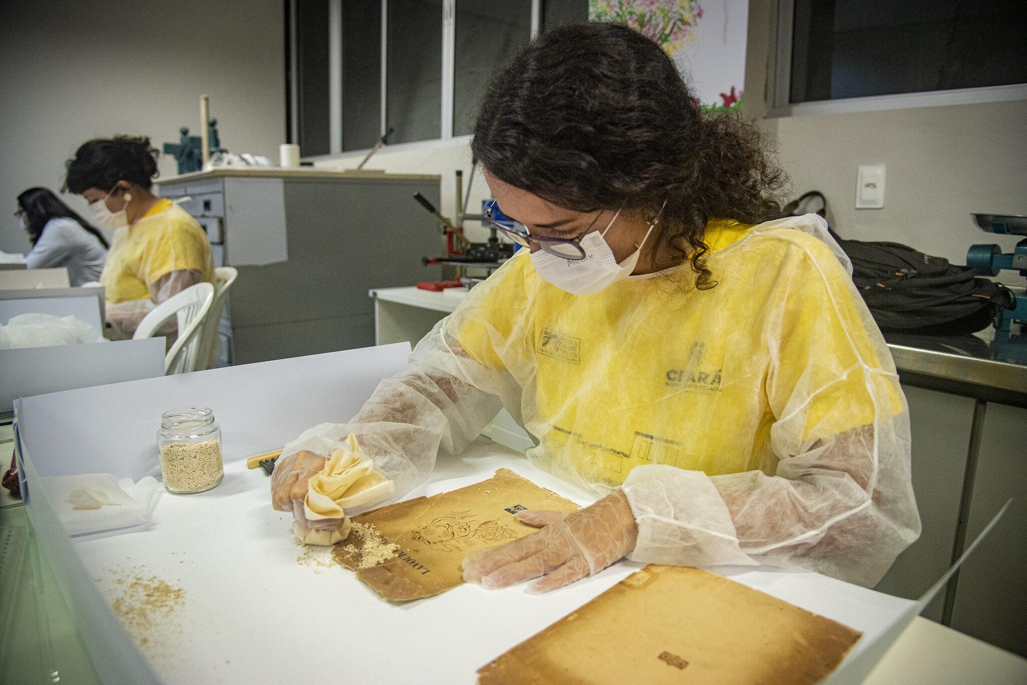 Menina usando jaleco, máscara e luva manipula página de obra rara para fazer restauração