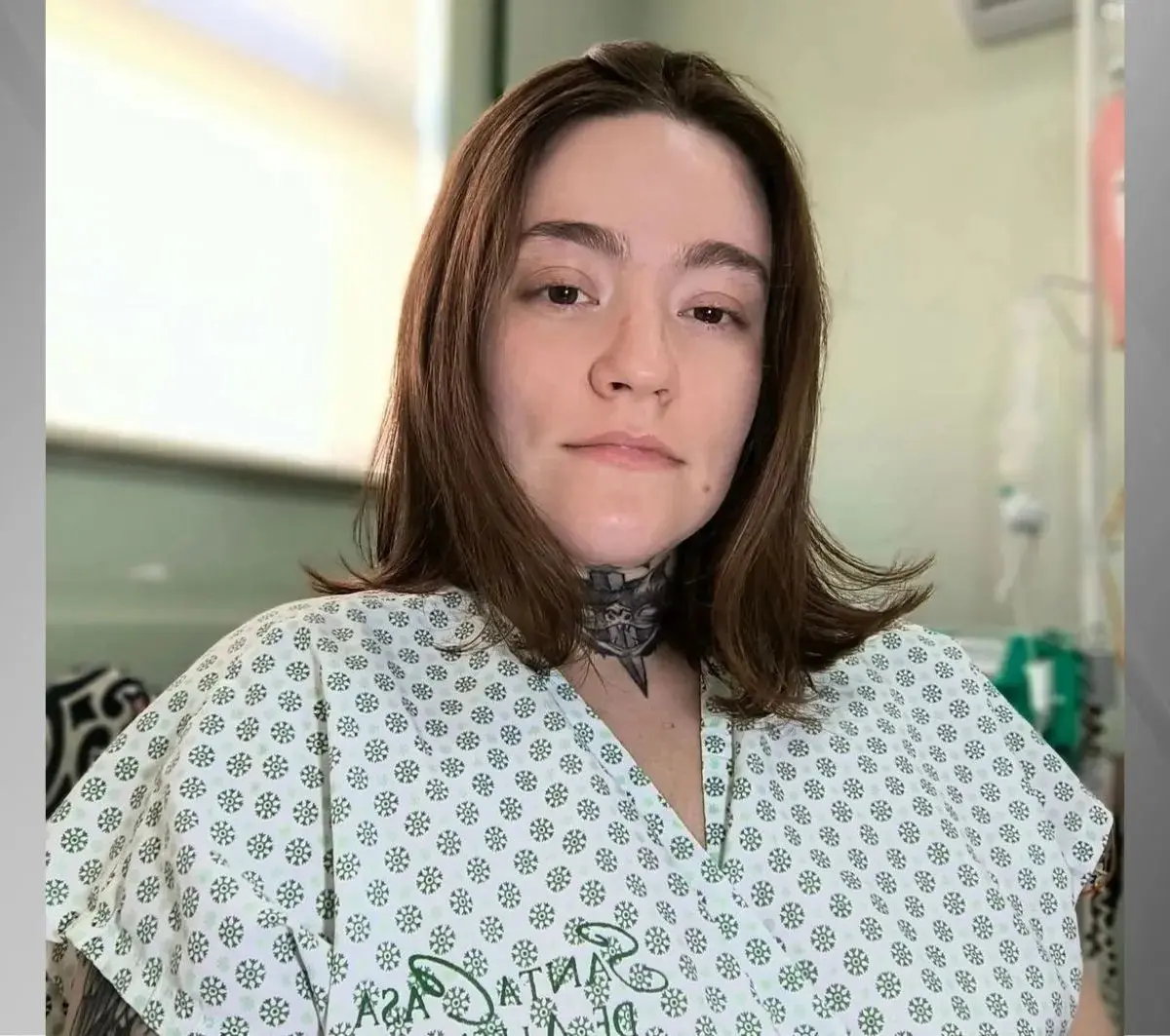 Carolina Arruda na sala de hospital com roupa de hospital e tatuagem no pescoço, sorrindo suavemente para a câmera.