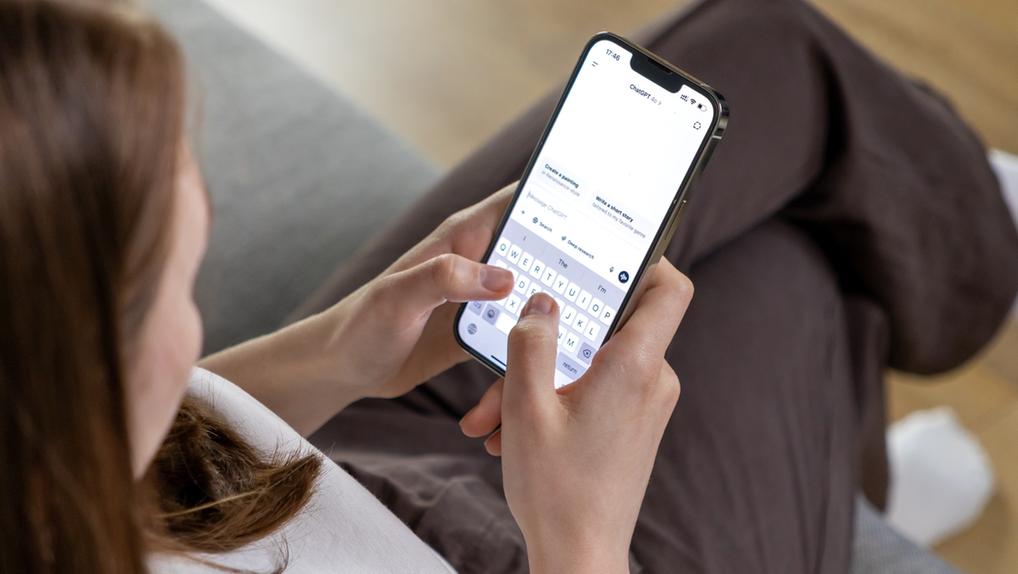 Imagem mostra mulher sentada em sofá, com o celular na mão. Na tela, está aberto o aplicativo do ChatGPT.