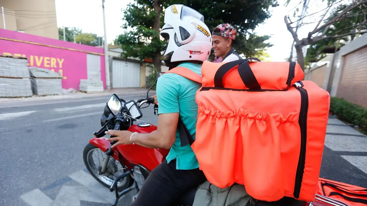 Foto que contém entregador de aplicativo, com capacete branco e camisa azul, em cima da mota com mochila laranja. Ele está parado