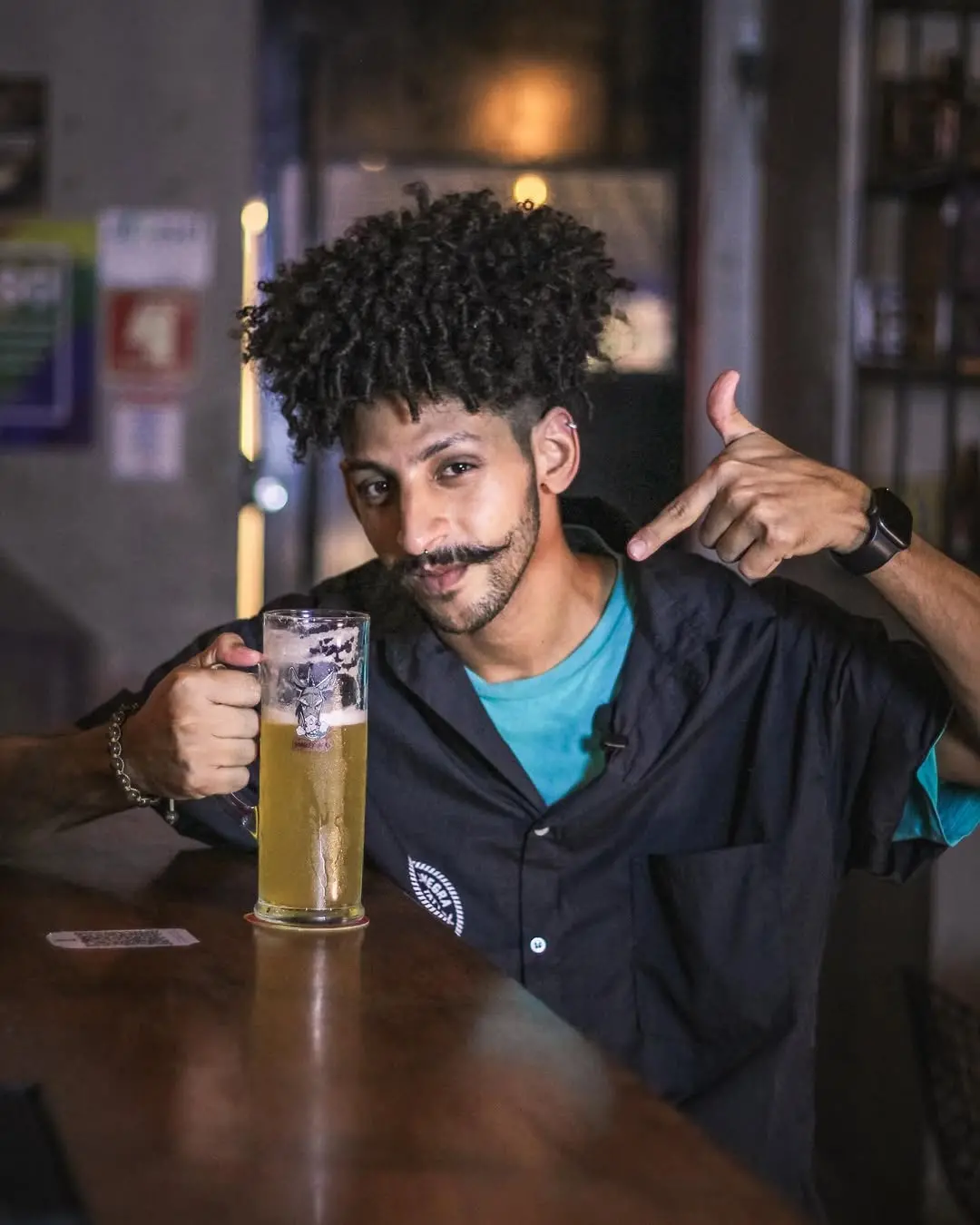 Foto que contém homem vestido de preto com expressão positiva enquanto segura caneco de chope