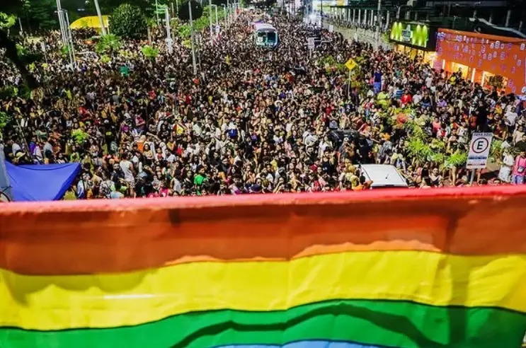 Multidão ocupa avenida à noite durante a Parada do Orgulho LGBTI+, com bandeira do arco-íris em destaque na frente da imagem