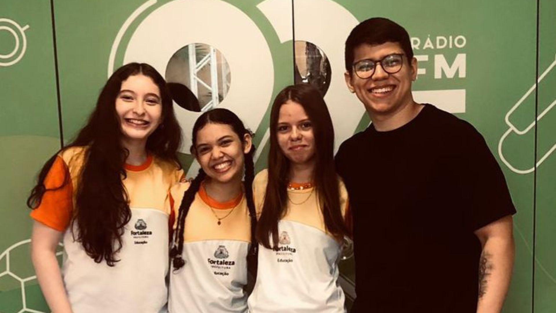 A imagem mostra três adolescentes abraçadas e um professor. Elas usam farda da rede pública municipal de Fortaleza