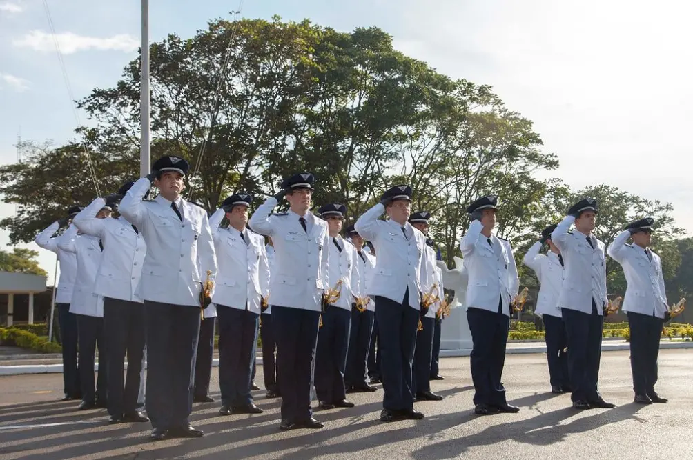 Foto de homens concludentes com fardamento branco da Aeronáutica, prestando continência, em cerimônia de formatura após curso de graduação no ITA. Ao fundo, árvores e sol