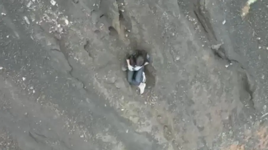 Imagem aérea mostra brasileira Juliana sentada na região vulcânica após cair em trilha de vulcão e equipes de resgatam tentam salvar mulher