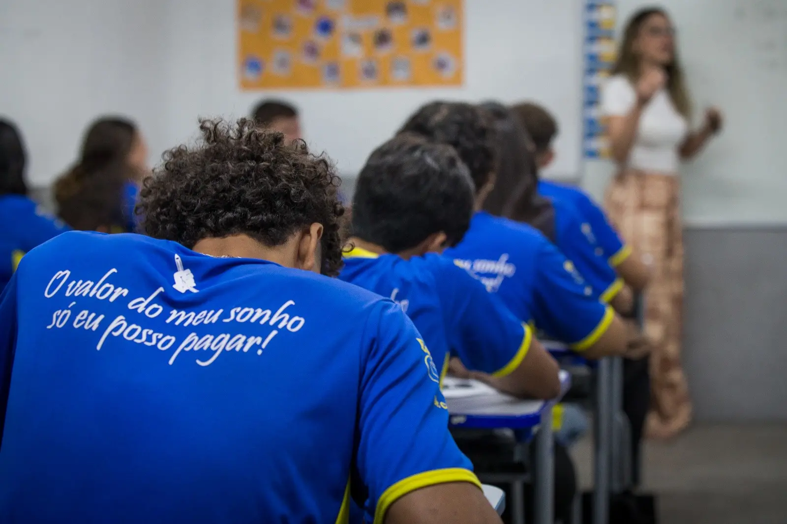 Estudantes assistem aula em sala de aula; na camisa de um deles lê-se: 