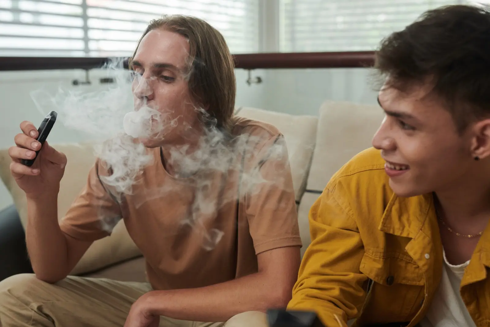 Imagem mostra dois jovens sentados em um sofá; um exala uma grande nuvem de vapor de um cigarro eletrônico, enquanto o outro observa sorrindo.