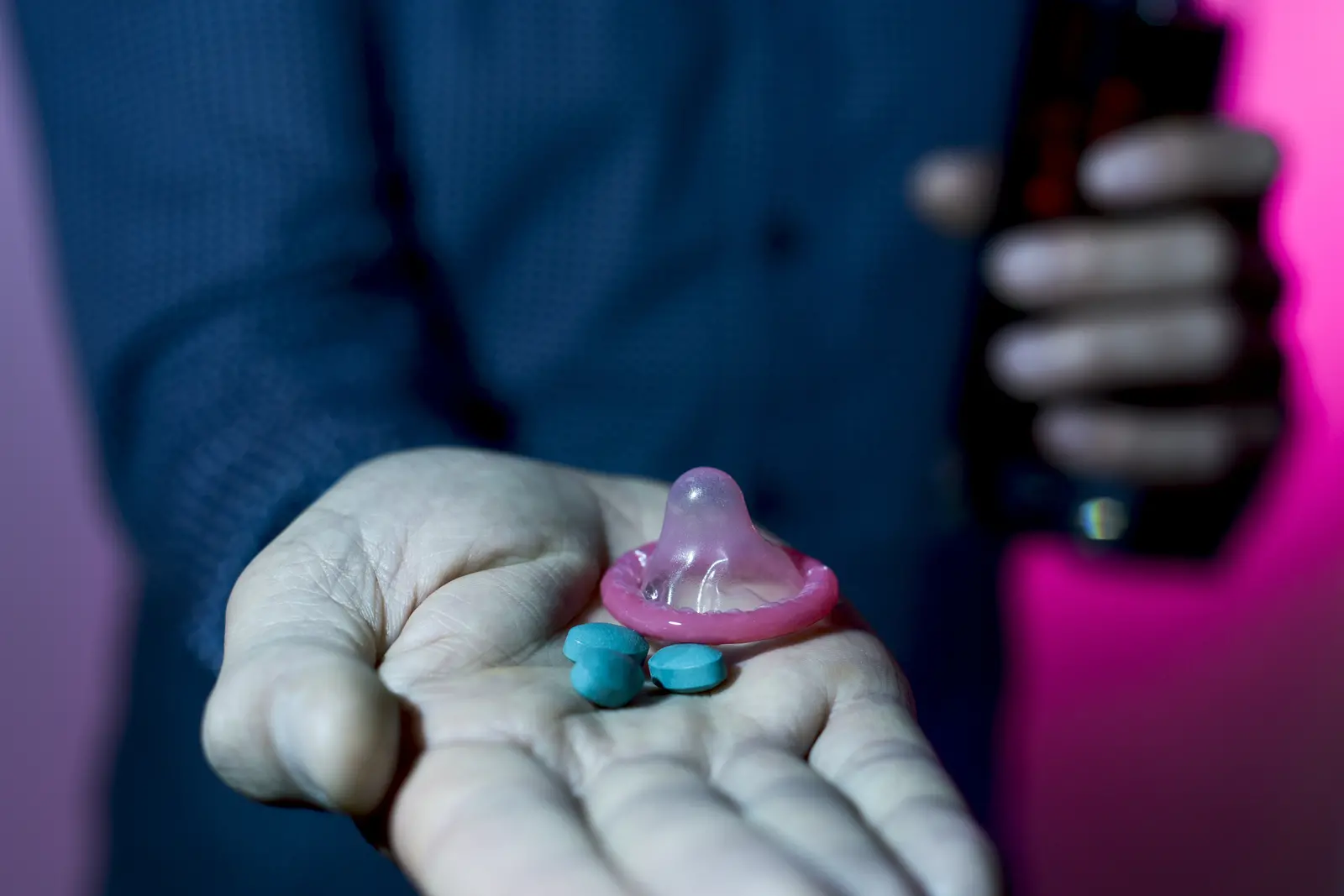 Uma mão aberta segura uma camisinha rosa e dois comprimidos azuis, com uma pessoa embaçada ao fundo.