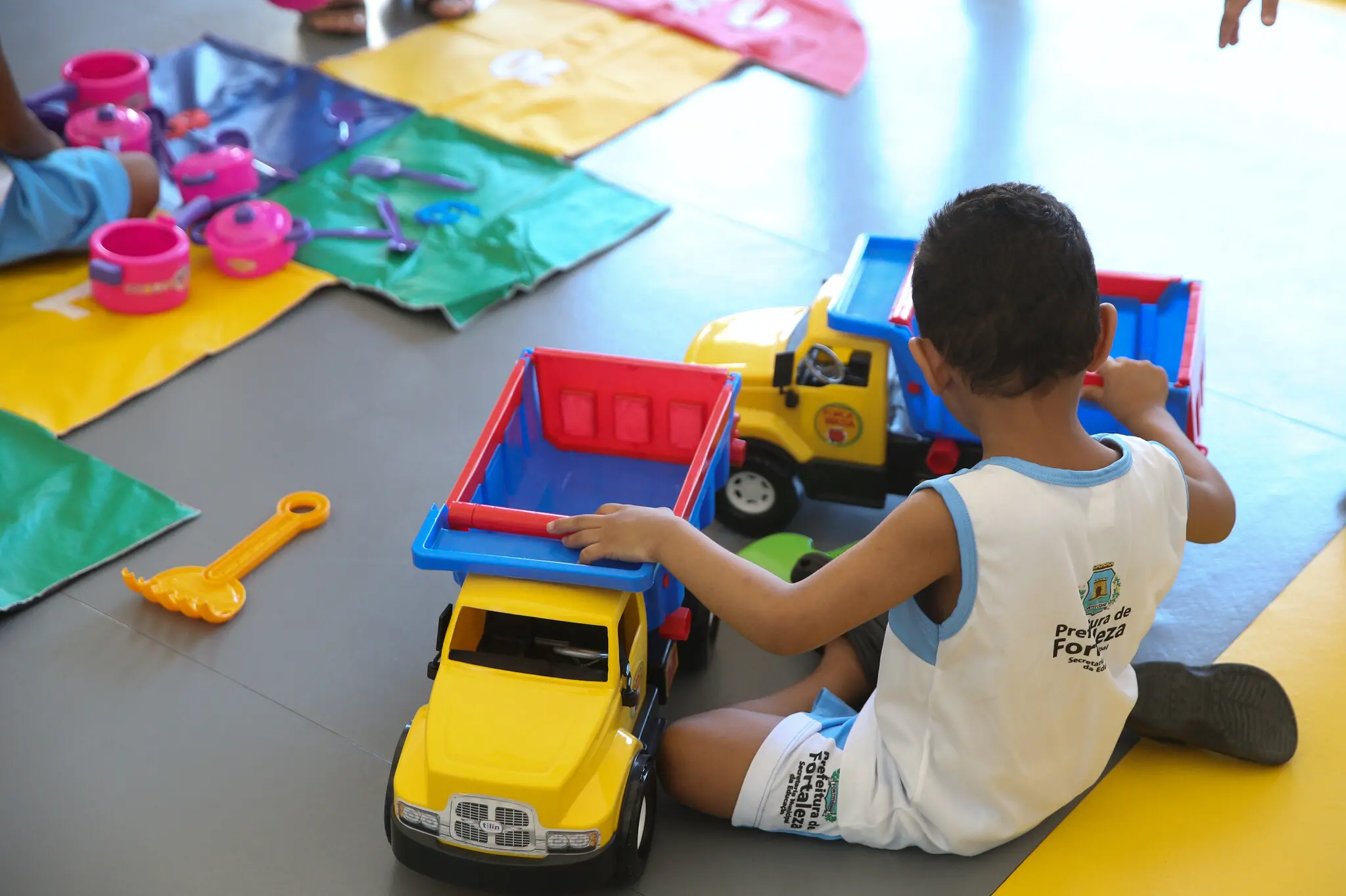 Criança com farda da prefeitura de Fortaleza, sentada no chão, de costas, com caminhões de brinquedo