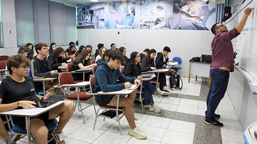 A foto mostra uma aula em uma sala de cursinho, onde um professor de pele morena, camisa vinho e calça jeans escura, escreve em um quadro branco à direita. Ele usa óculos e tem barba. A sala tem fileiras de carteiras, onde alunos jovens, em sua maioria de pele clara, estão sentados. Muitos estão de cabeça baixa, concentrados em seus cadernos ou telas de notebook. Ao fundo, uma parede decorada com imagens de profissionais da saúde. O piso é branco com detalhes em cinza.