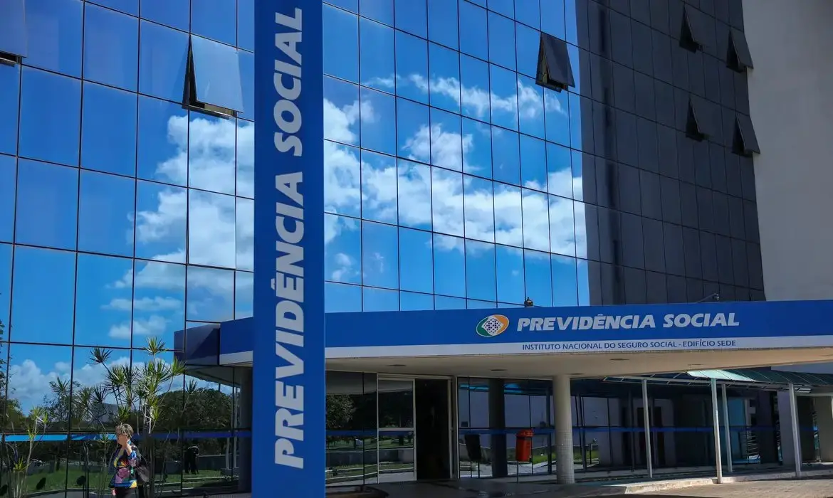 Presidente Lula assina decreto que altera regras do BPC; fachada do prédio do INSS com o logo da Previdência Social refletindo o céu azul com nuvens em estrutura de vidro espelhado.