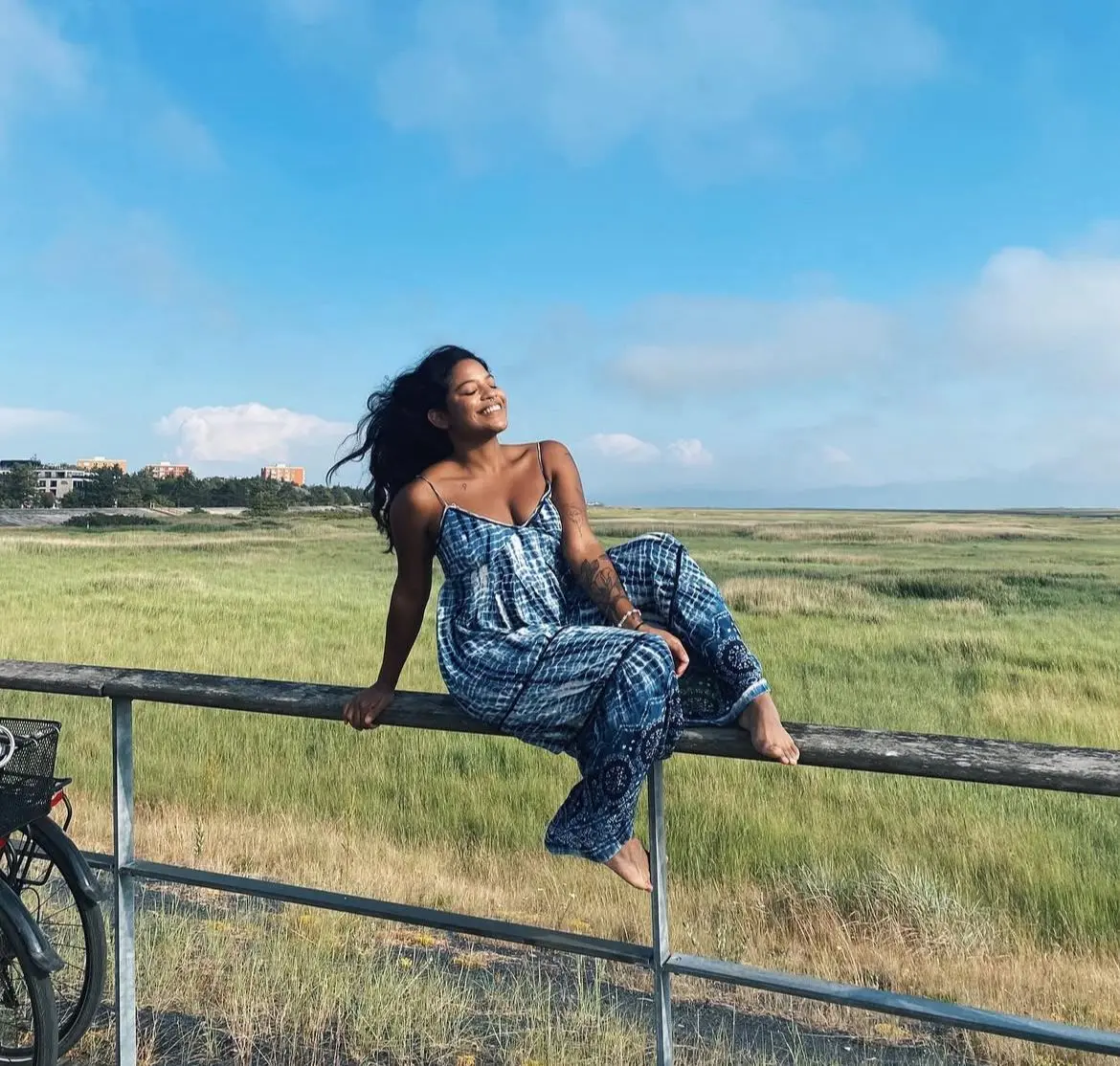 Juliana Marins, sorrindo sentada em uma grade, em meio a uma paisagem aberta de campo verde e céu azul
