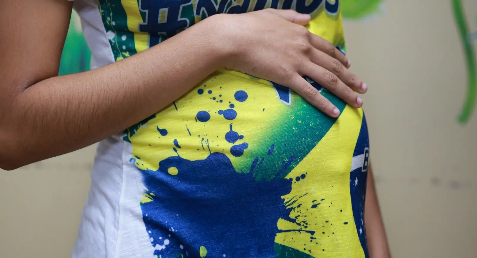 Foto de mulher grávida com braço sobre a barriga. Ela veste uma blusa com cores da bandeira do Brasil, em amarelo, azul e verde