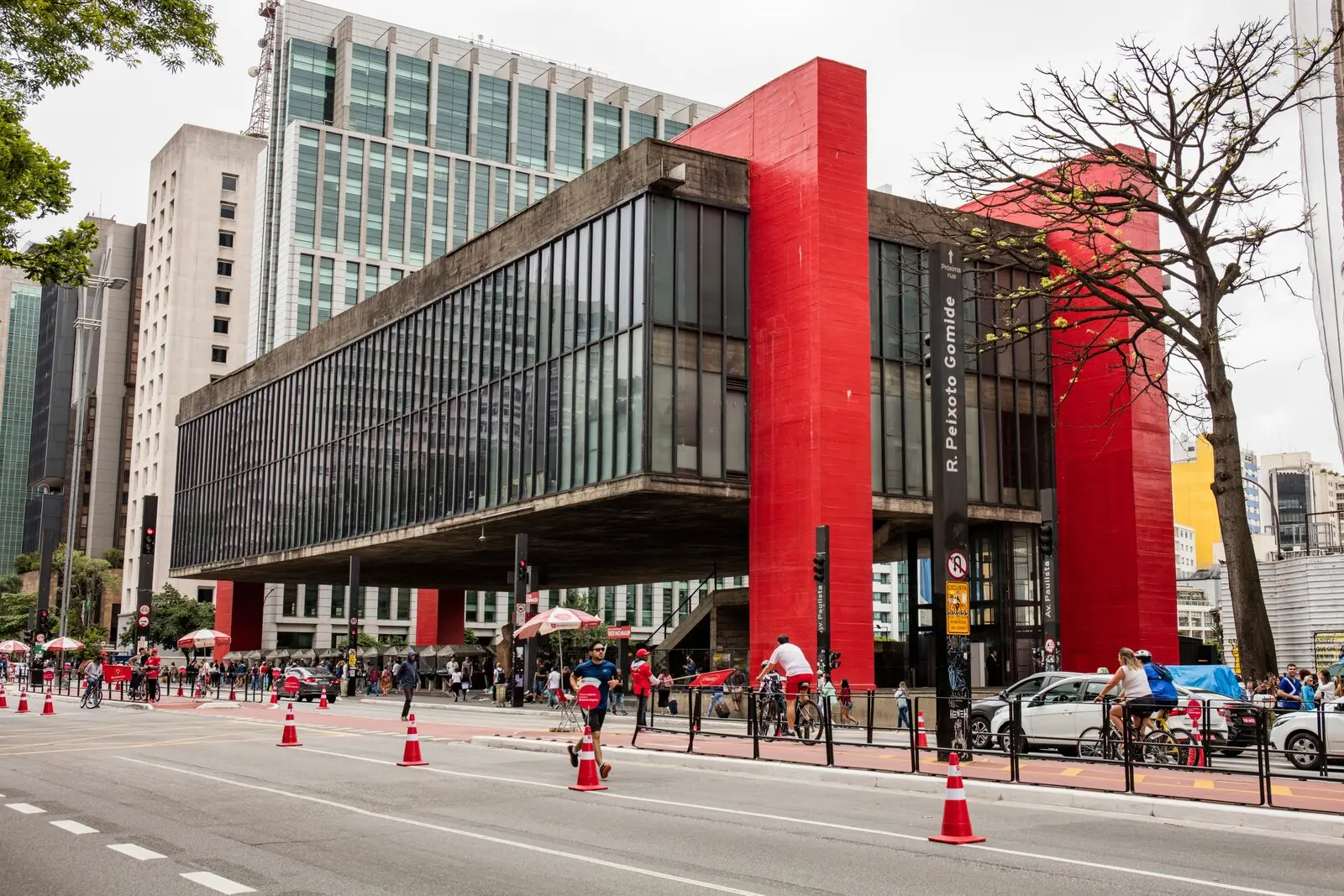 A imagem mostra uma rua movimentada com pessoas e carros, ladeada pelo MAPS, um edifício moderno de arquitetura marcante em vermelho e preto, em São Paulo, e outros prédios altos ao fundo.