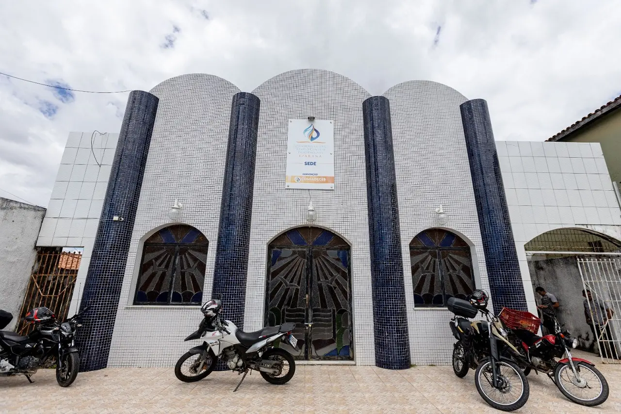 Foto da fachada da Assembleia de Deus Templo Central de Iparana, em azulejos brancos com quatro fileiras verticais azuis e placa de identificação. No pátio em frente, há quatro motos estacionadas