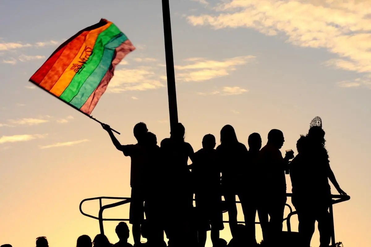 Imagem mostra silhuetas de um grupo de pessoas em cima de um trio, durante a Parada pela Diversidade Sexual do Ceará. Uma das pessoas empunha uma bandeira com as cores LGBTI+. Ao fundo, o céu com luz de pôr do sol.