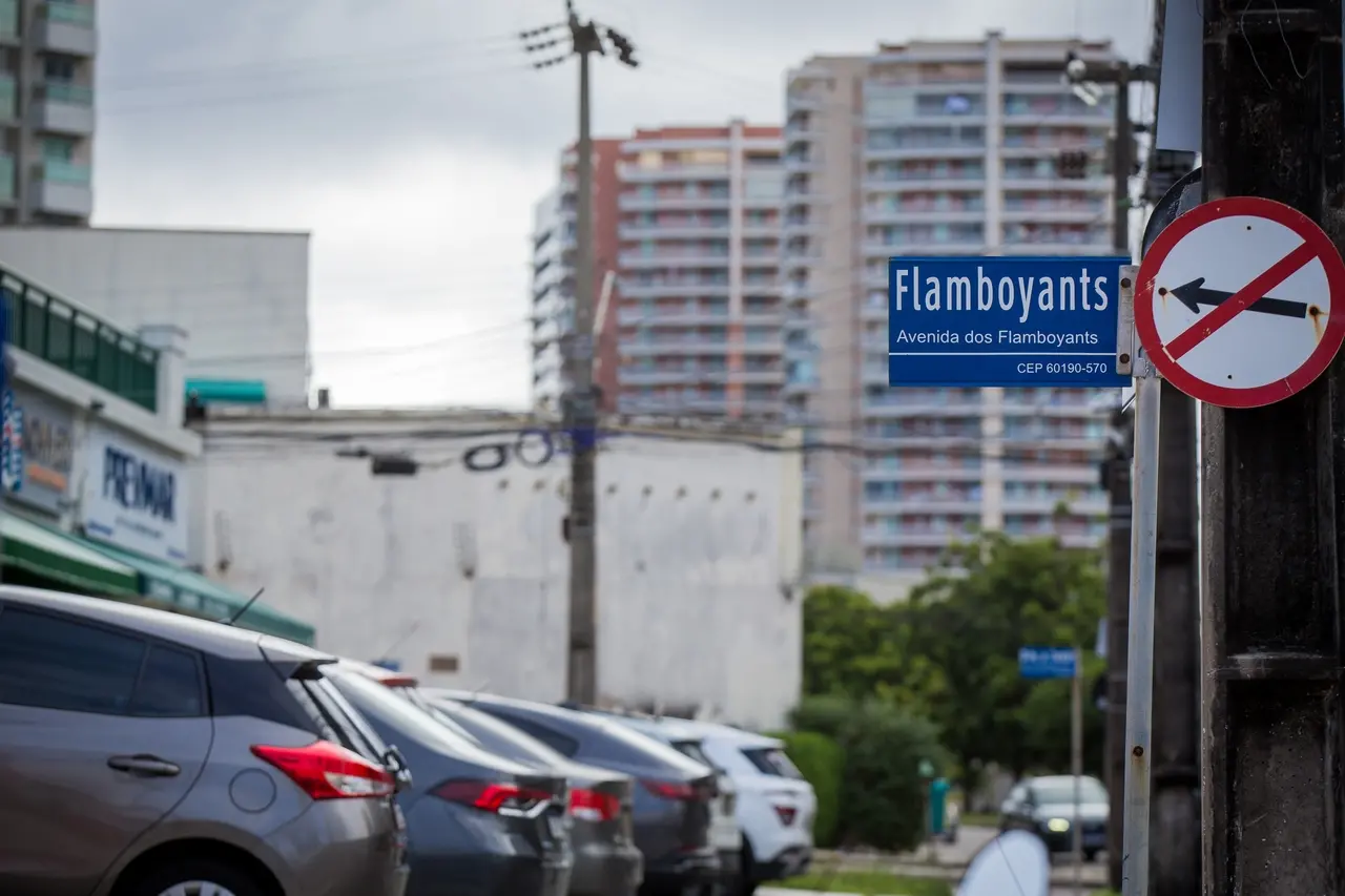Imagem mostra a placa de uma rua. Ao fundo, carros estacionados e um prédio