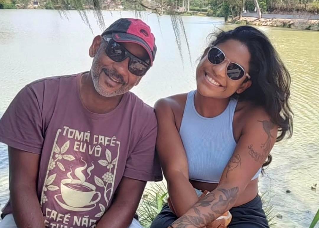 Manoel Marins e Juliana Marins sentados lado a lado na beira de um lago. Eles estão sorrindo e de óculos escuros