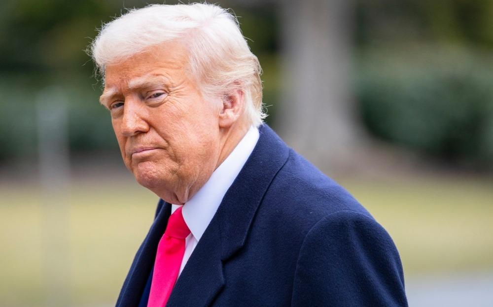 Donald Trump é um homem idoso branco de cabelos grisalhos penteados para o lado. Na foto, ele está de terno azul, camisa social branca e gravata vermelha