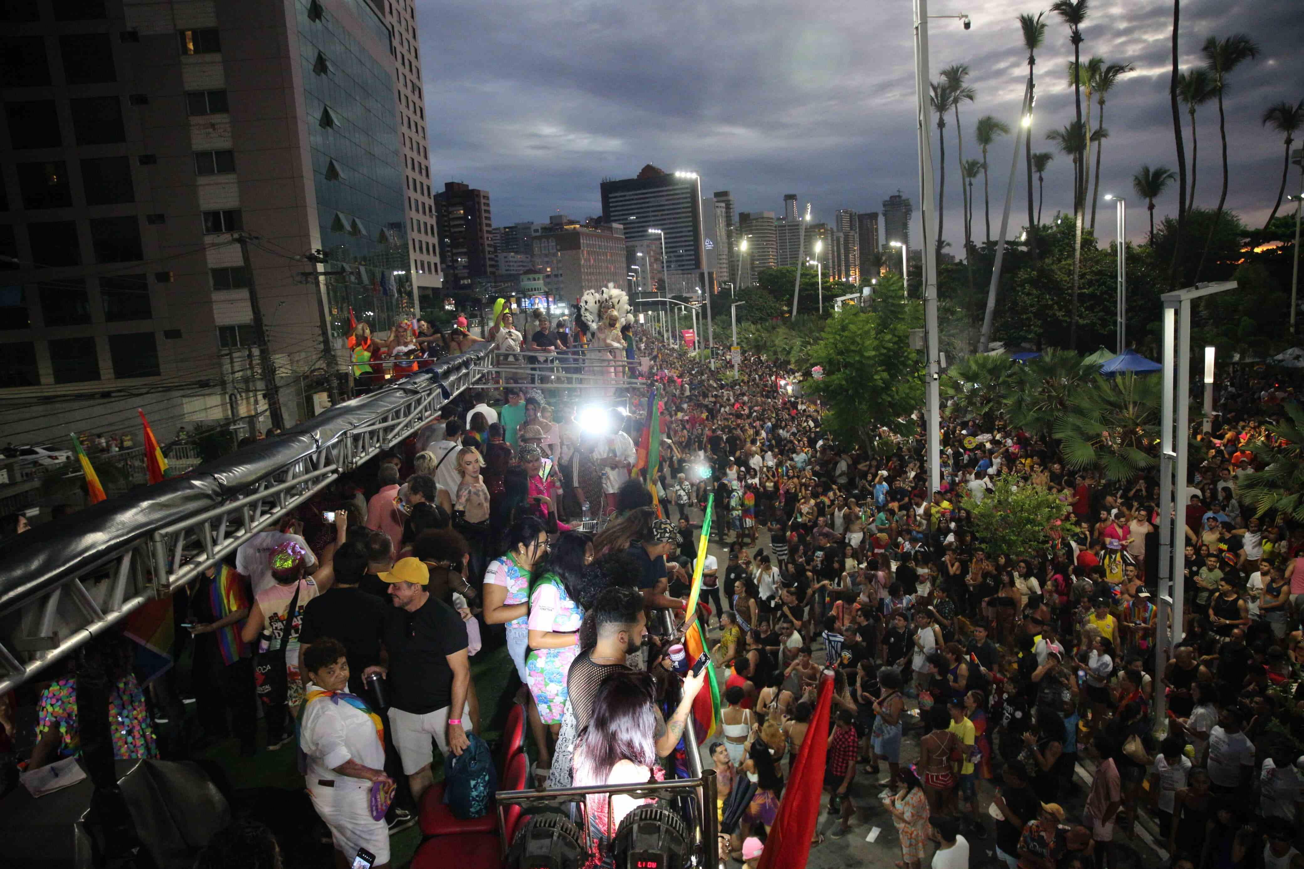 Imagem de uma grande manifestação ou evento de rua durante o entardecer, com milhares de pessoas reunidas em uma avenida entre prédios altos e palmeiras iluminadas por postes. No canto esquerdo da imagem, há um trio elétrico lotado de participantes, muitos com roupas coloridas, fantasias e bandeiras, incluindo bandeiras do arco-íris, símbolo do movimento LGBTQIA+. As pessoas na rua seguem o trio elétrico, compondo uma multidão que se estende até o horizonte. O céu está parcialmente nublado, e as luzes da cidade já estão acesas.