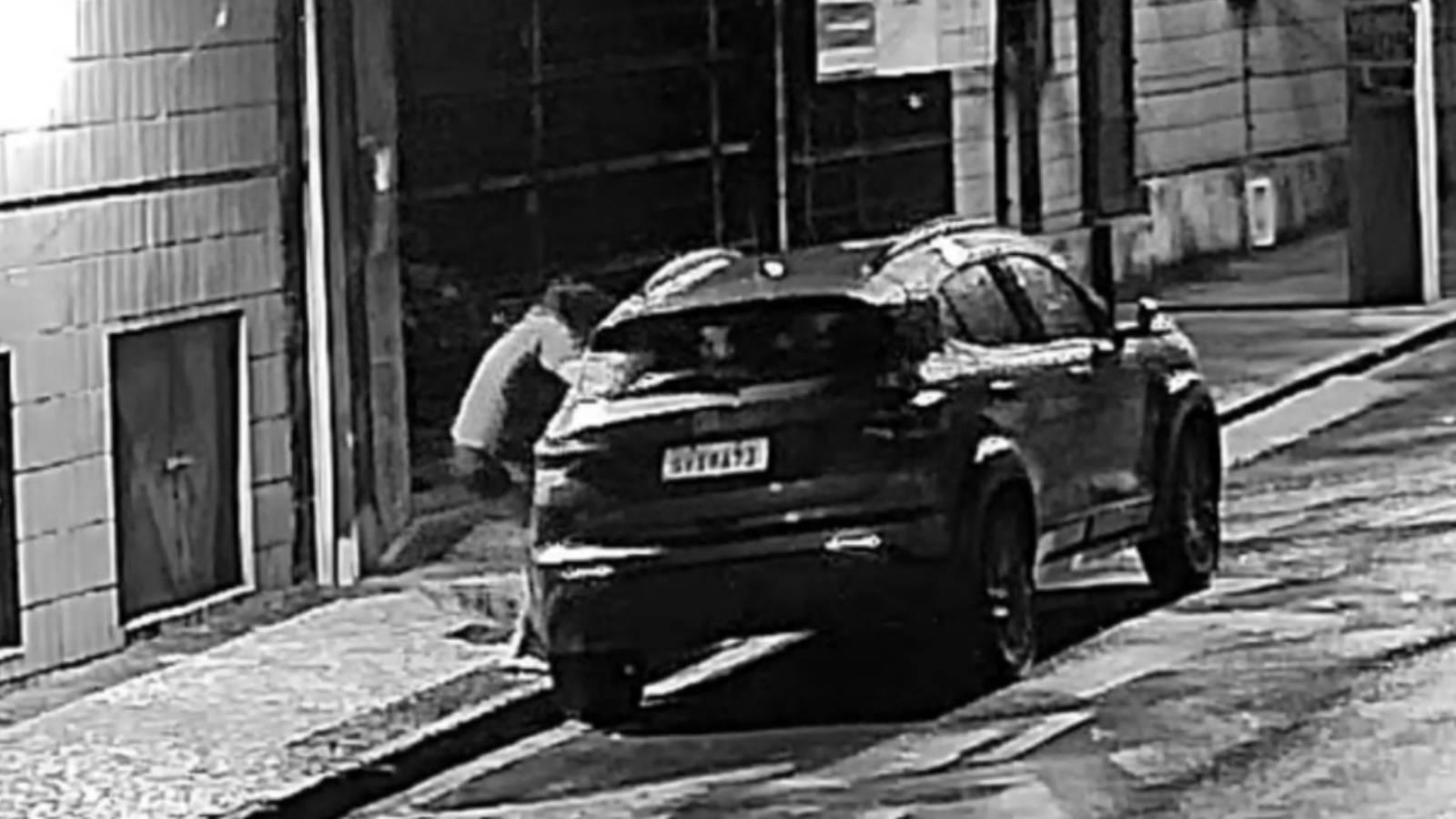 Imagem de uma rua com um carro preto estacionado, em rua do bairro Meireles, zona nobre de Fortaleza, e uma mulher furtando objetos do seu interior após quebrar a janela. Cena noturna em ambiente urbano.