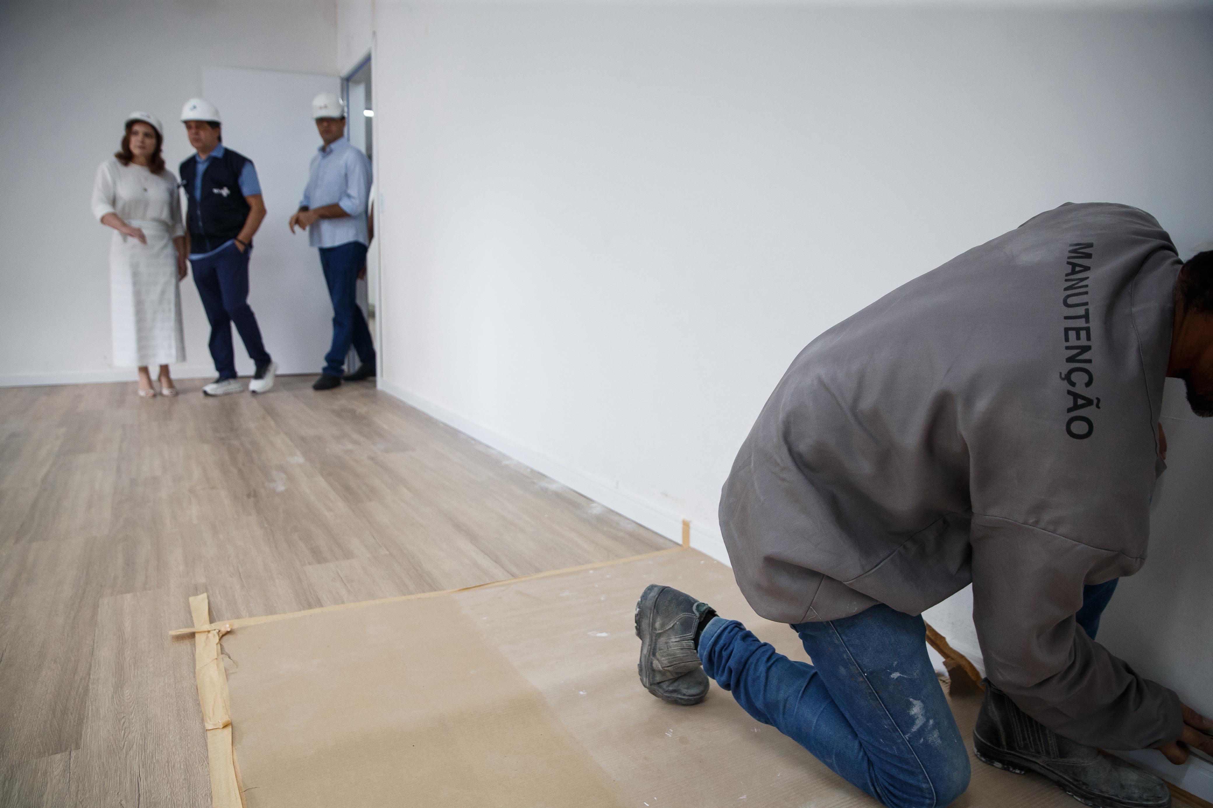 Imagem mostra, no canto inferior direito, um homem pintando uma parede de branco, no Centro de Diagnóstico Girassol, em Fortaleza; No canto superior esquerdo, em segundo plano, aparecem três pessoas visitando a sala em reforma: Cristiane Leitão, primeira-dama de Fortaleza; Evandro Leitão, prefeito da cidade; e um engenheiro.