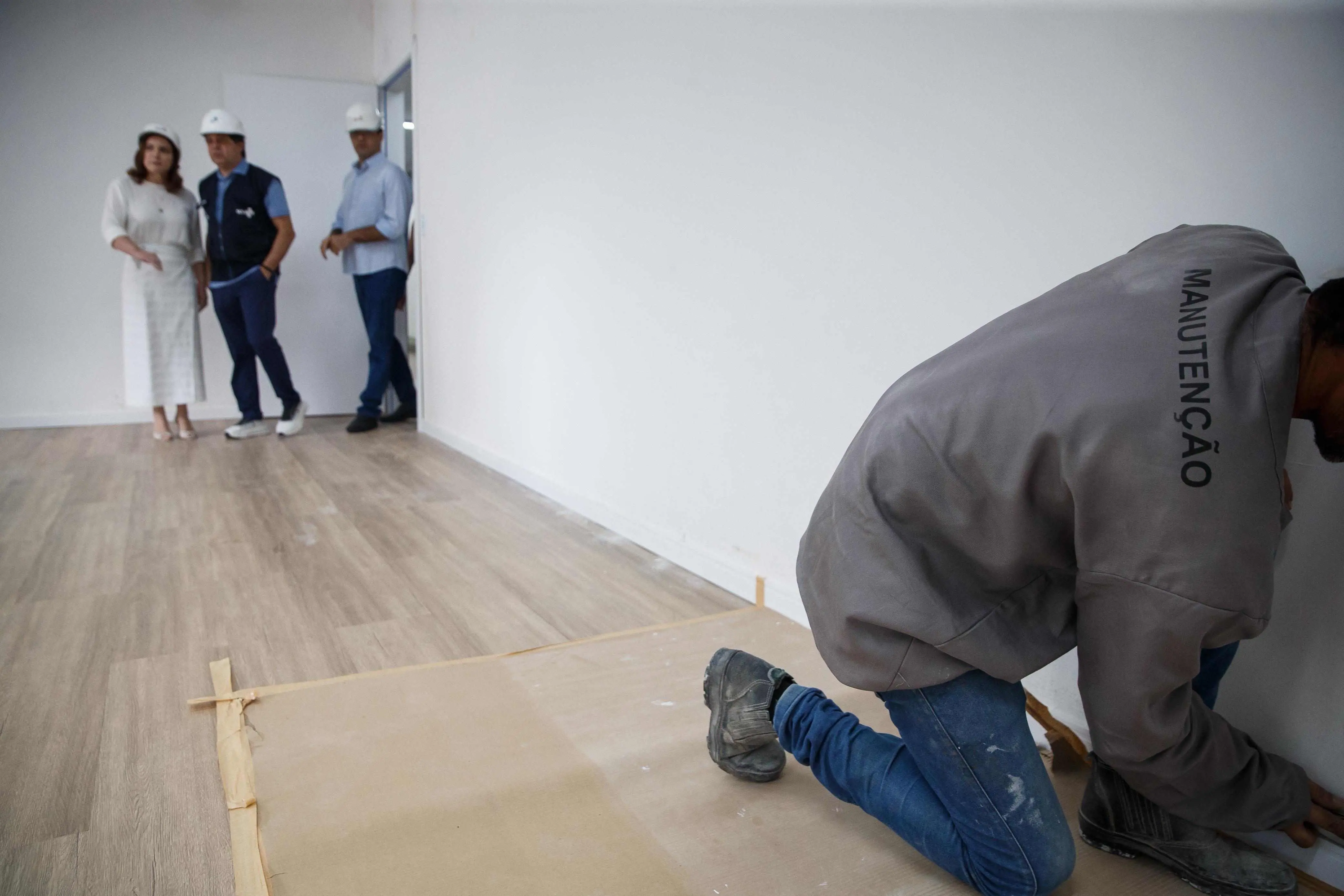Imagem mostra, no canto inferior direito, um homem pintando uma parede de branco, no Centro de Diagnóstico Girassol, em Fortaleza; No canto superior esquerdo, em segundo plano, aparecem três pessoas visitando a sala em reforma: Cristiane Leitão, primeira-dama de Fortaleza; Evandro Leitão, prefeito da cidade; e um engenheiro.
