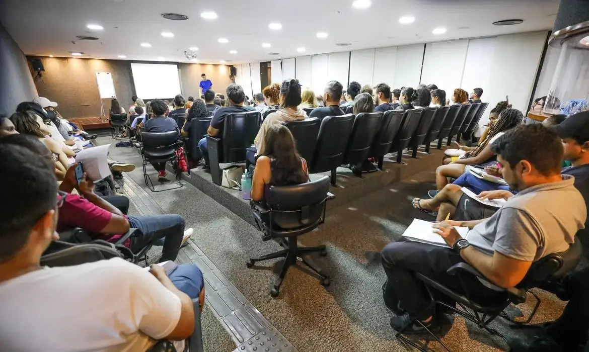 Pessoas assistindo a uma palestra ou apresentação em uma sala de conferências com muitos assentos e uma pessoa falando na frente do grupo