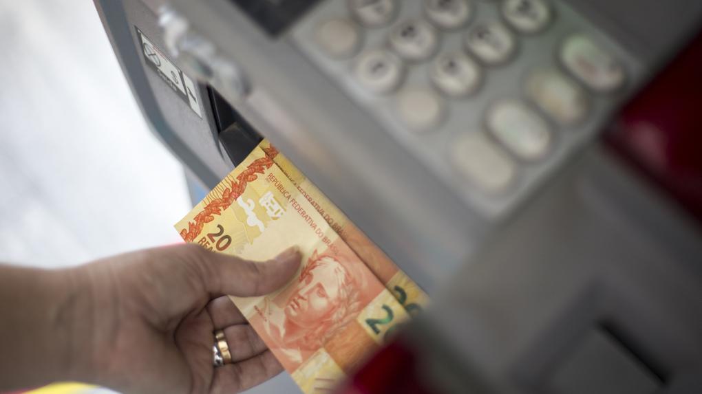 Pessoa sacando dinheiro de uma máquina de caixas eletrônicos, com notas de R$ 20 em mãos, ilustrando pagamento da primeira parcela do 13º salários aos servidores do Governo do Estado e da Prefeitura de Fortaleza