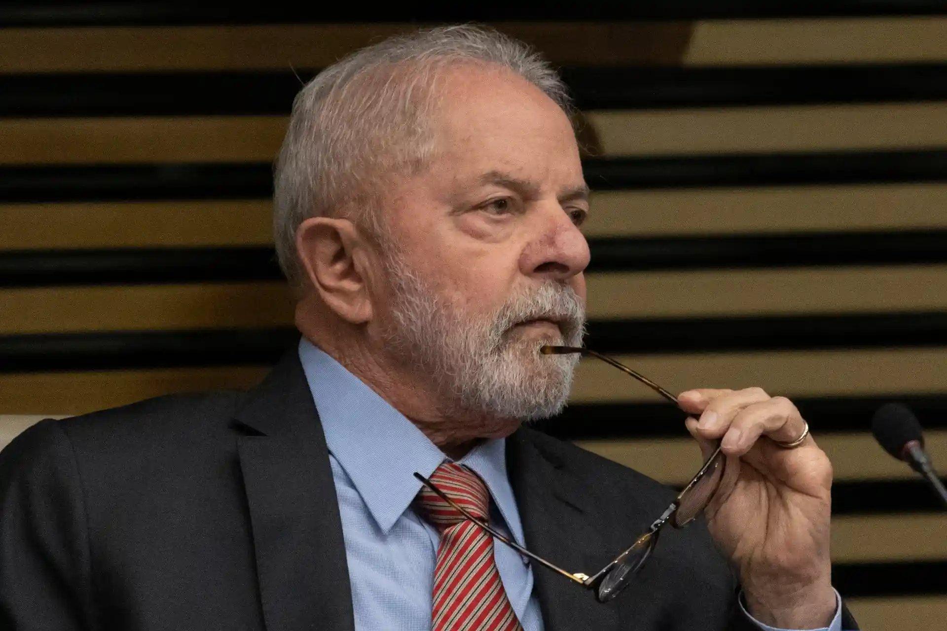 Presidente Lula em ambiente interno com parede de madeira, usando terno e gravata e segurando óculos de grau, em momento de reflexão. Governo Lula aciona STF para tentar restabelecer decreto que aumenta alíquotas do IOF e reforça arrecadação.