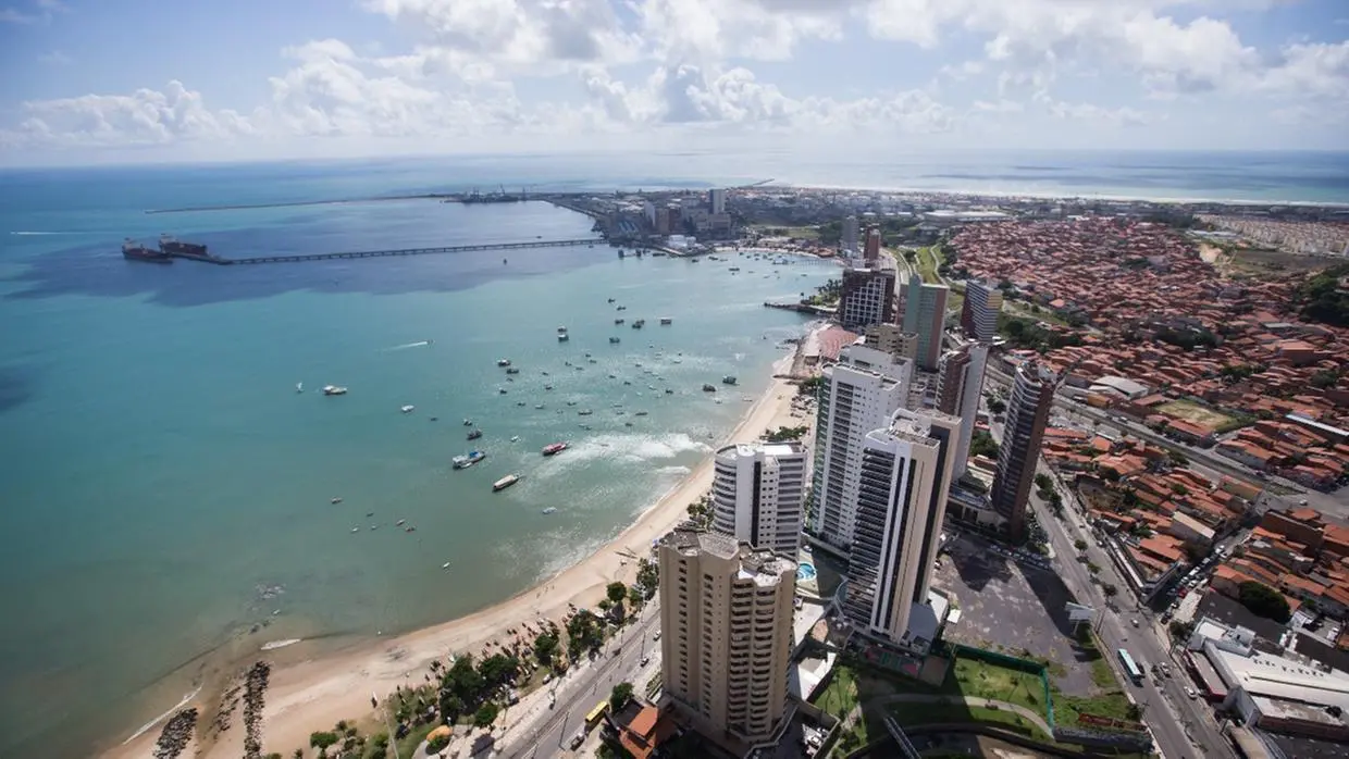 Foto aérea da orla de Fortaleza, no Ceará, mostrando prédios altos à beira-mar, barcos ancorados no mar calmo e um píer que se estende até navios ao fundo. Ao lado direito, vê-se uma área urbana com muitas casas de telhado vermelho.