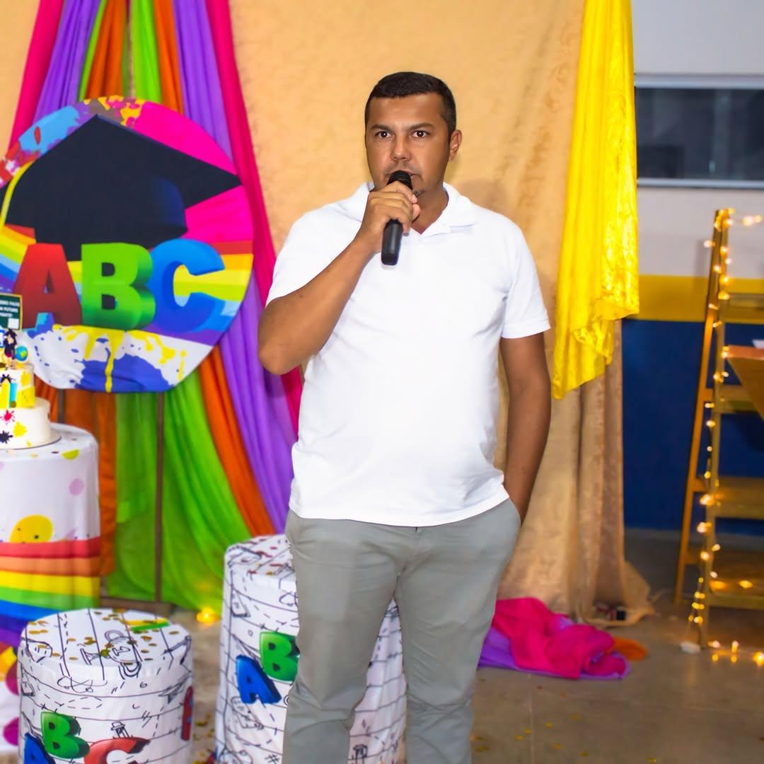 Imagem mostra Francisco Alberto em pé segurando um microfone, aparentemente discursando em um evento. Ele veste camisa polo branca e calça cinza. Ao fundo, há uma decoração colorida com tecidos em tons de rosa, roxo, verde e laranja, além de um grande círculo com as letras 