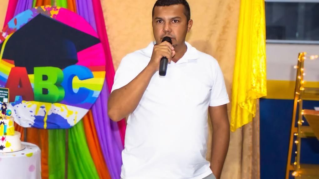 Imagem mostra Francisco Alberto em pé segurando um microfone, aparentemente discursando em um evento. Ele veste camisa polo branca e calça cinza. Ao fundo, há uma decoração colorida com tecidos em tons de rosa, roxo, verde e laranja, além de um grande círculo com as letras