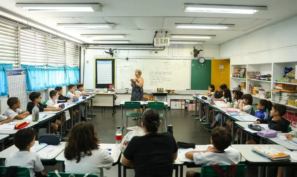 Sala de aula com crianças assistindo à professora na frente, em um ambiente escolar bem iluminado e organizado, com quadro negro, livros e materiais escolares. Confira como solicitar a isenção da taxa de inscrição da Prova Nacional do Docente.