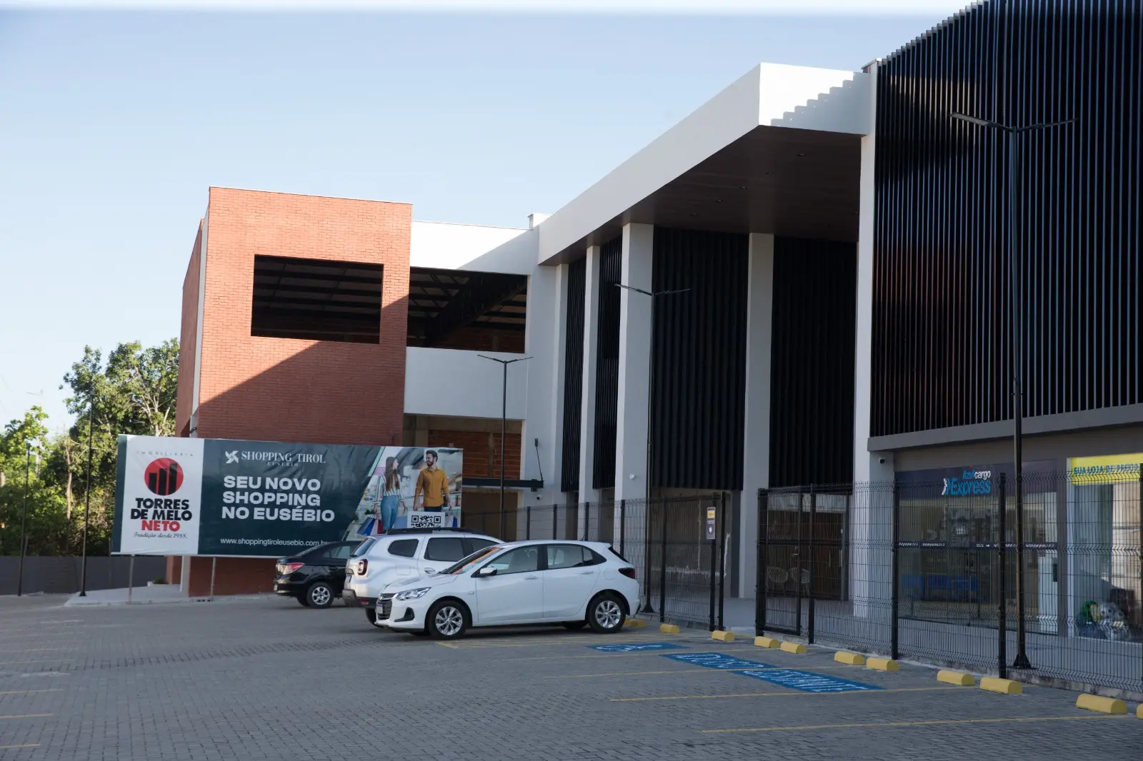Foto que contém um pátio comercial no Eusébio. Na área externa, estão carros estacionados. Algumas lojas ainda estão fechadas