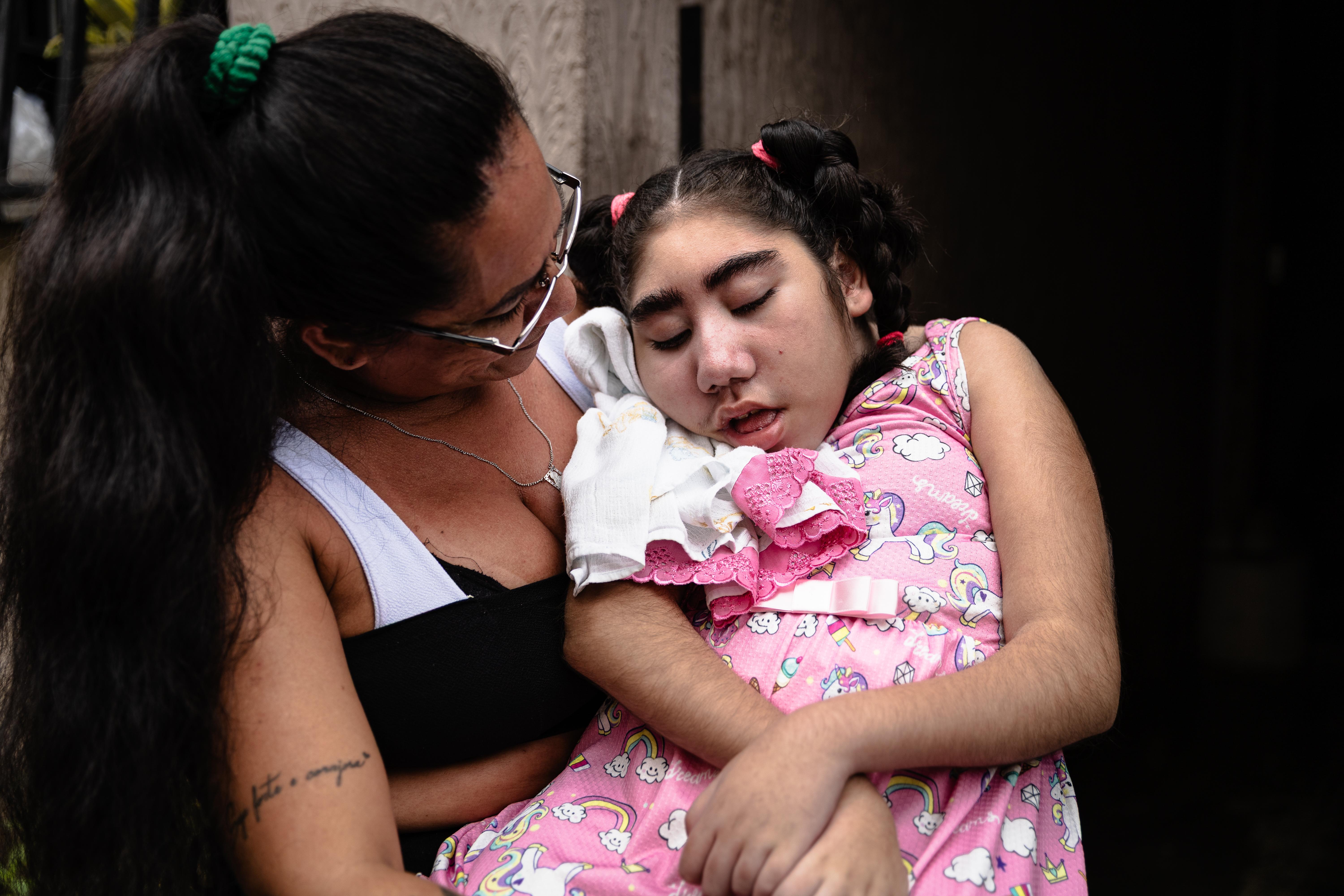 A foto mostra uma mãe e uma criança estão sentadas. A mulher usa uma blusa preta com alças brancas e a criança, deitada em seu colo, está com cabelo em duas tranças e um vestido rosa