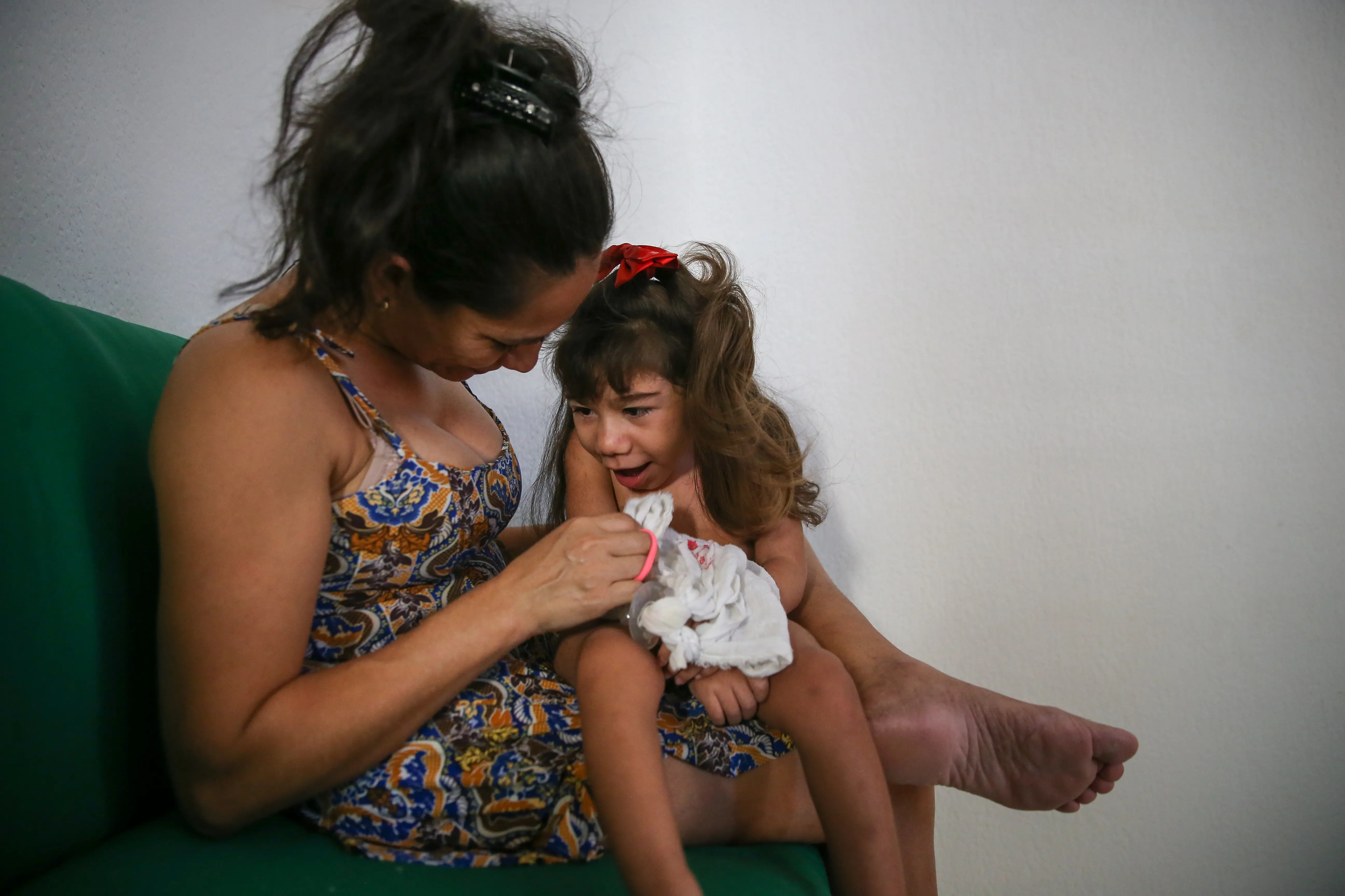 A foto mostra uma mãe e uma criança estão sentadas em um sofá verde. A mulher usa um vestido estampado e tem o cabelo escuro preso. A criança, com cabelo comprido e um laço vermelho, está sentada no colo dela segurando um pedaço de tecido