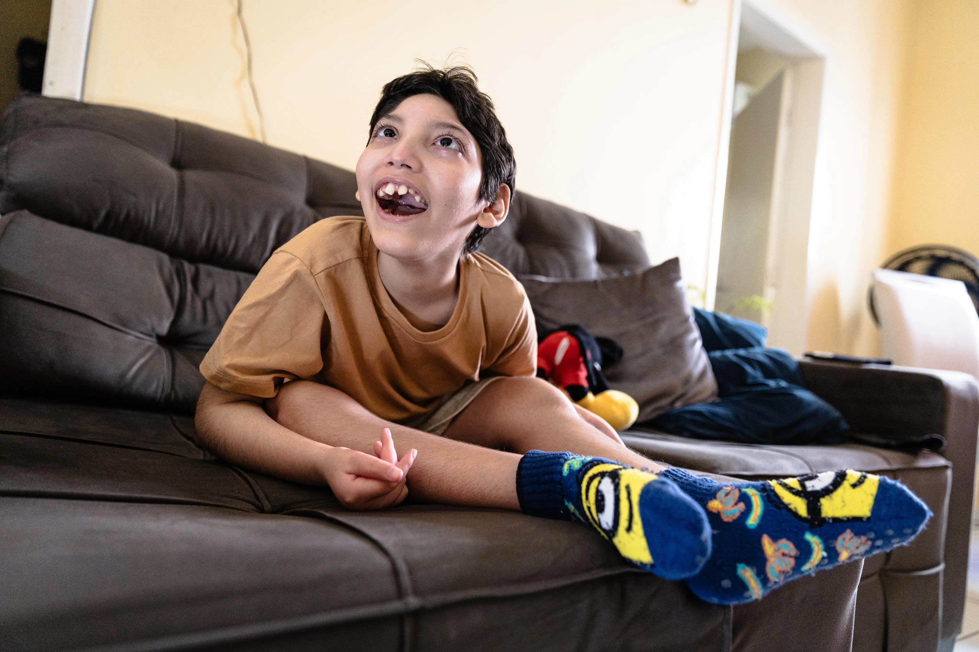 A foto mostra uma criança sentada em um sofá cinza, vestindo uma camisa marrom, shorts e meias azuis com personagens Minions. Há almofadas e brinquedos ao lado. Ao fundo, vê-se um cômodo claro com uma porta aberta para outra área da casa