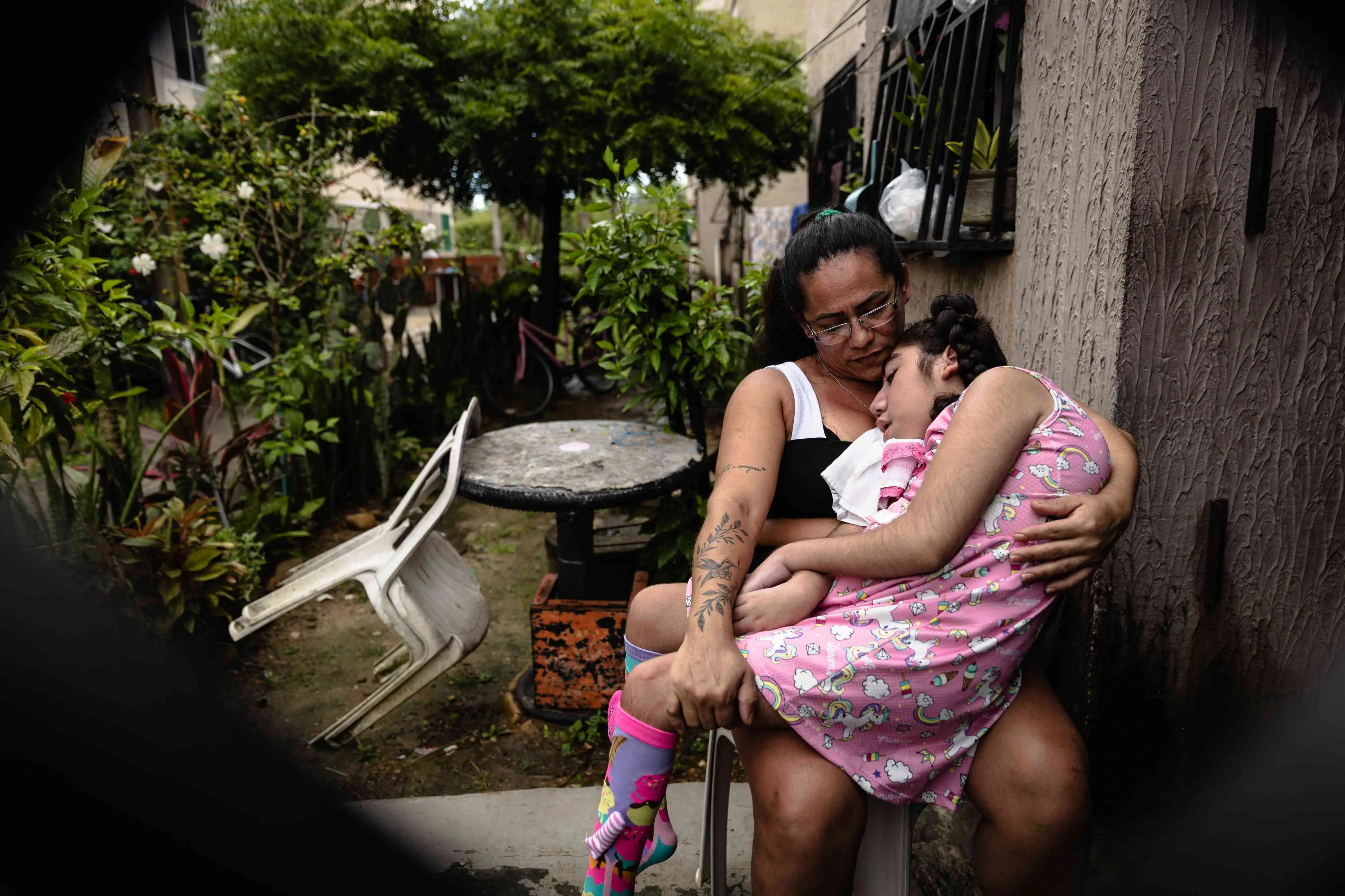 A foto mostra duas pessoas sentadas em uma cadeira de plástico fora de um prédio; a mãe segura a filha no colo, ninando ela. A criança usa meias coloridas com estampas de doces. Há vasos de plantas, cadeira, mesa e janelas com grades ao fundo.