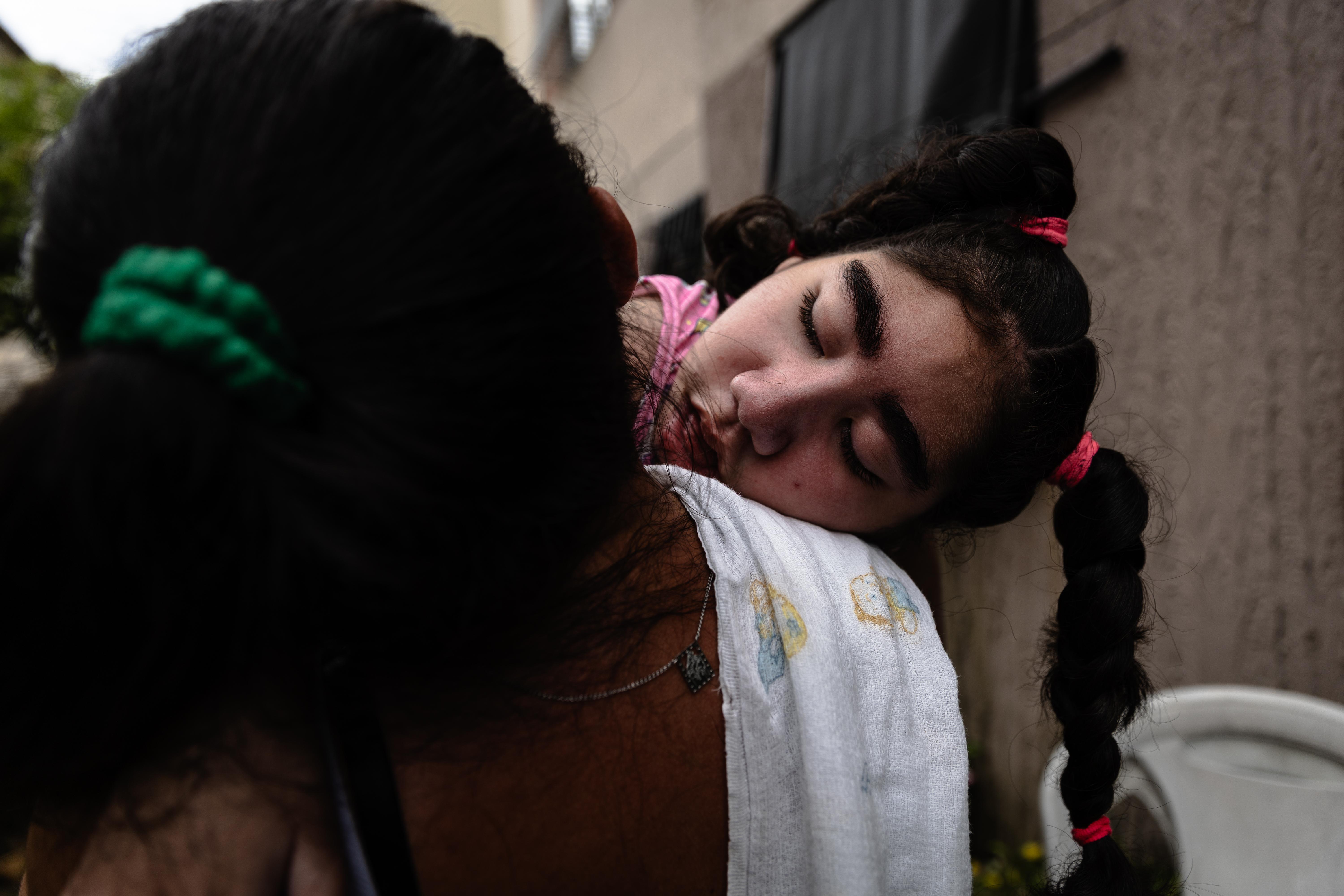 A foto mostra a mãe de costas com cabelo preso por um elástico segurando a filha. A criança tem tranças com elásticos rosas e usa uma toalha branca sobre o ombro. Ao fundo, há prédios e vegetação
