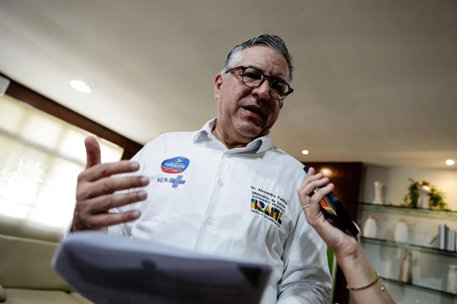A imagem mostra o ministro da Saúde, Alexandre Padilha, usando óculos e camisa branca, falando e gesticulando com as mãos. Na camisa, há o logo do Governo Federal e do SUS.