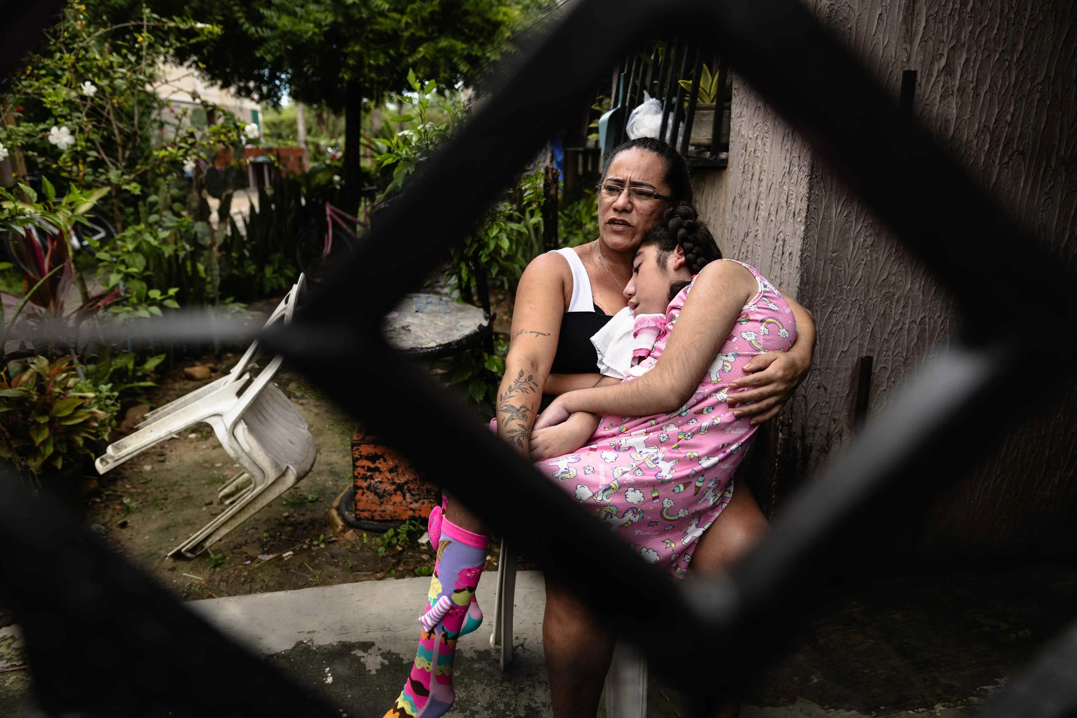 A foto mostra uma mãe sentada ao ar livre, segurando uma criança com vestido rosa estampado e meias rosas. A criança está com a cabeça apoiada no ombro da pessoa. Ao fundo, há plantas, uma cadeira plástica branca virada e uma mesa redonda.