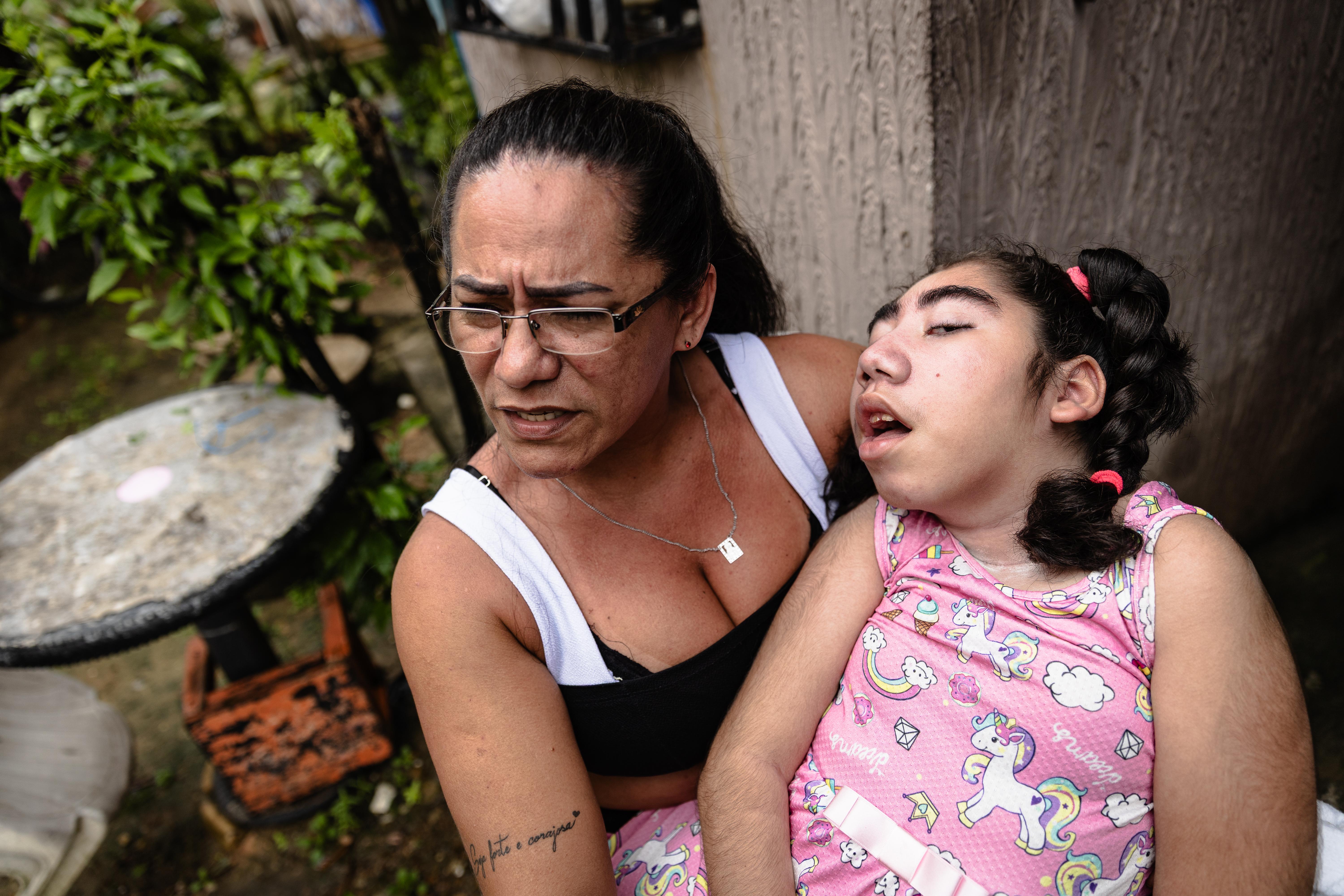A foto mostra uma mãe sentada ao ar livre, segurando uma criança com vestido rosa estampado e meias rosas. A criança está com a cabeça apoiada no ombro da pessoa. Ao fundo, há plantas, uma cadeira plástica branca virada e uma mesa redonda.