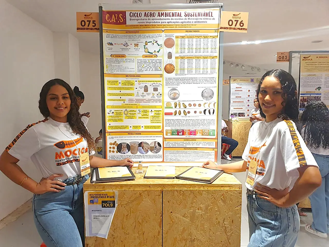 Duas jovens apresentadoras posando ao lado de um pôster sobre ciclo agro ambiental sustentável em uma feira de ciências escolar, com detalhes do projeto e outros pôsteres ao fundo.